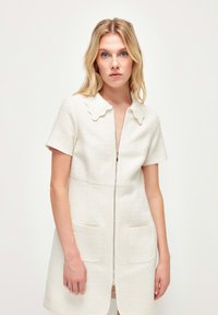 Vestido de color crema con mangas cortas, cuello con volantes, cremallera delantera y dos grandes bolsillos de parche; tejido texturizado.