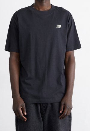 Person iført en sort New Balance t-shirt med korte ærmer og et lille hvidt logo på venstre bryst, kombineret med mørke bukser.