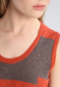 Stickad ärmlös topp med orange och mörkgrå färgblock. Rund halsringning med ribbade kanter. Mjuk, текстurerad tyg.