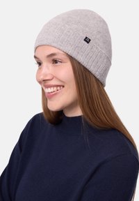 Berretto a maglia grigio chiaro con una texture a coste, dotato di un piccolo logo nero. Abbinato a un maglione blu scuro con collo alto.