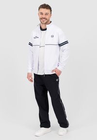 Witte zip-up jas met zwarte strepen en logo, gecombineerd met zwarte broek met zijstrepen en een logo, compleet met witte sneakers.