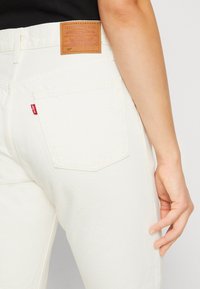 Jeans de denim blancos con una etiqueta de cuero marrón que dice "LEVI STRAUSS & CO." Costura roja en el bolsillo trasero en un patrón curvado.