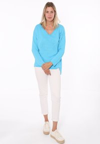 Felpa azzurra con scollo a V caratterizzata da un design in rilievo, abbinata a pantaloni bianchi slim-fit e scarpe da ginnastica bianche. Texture liscia.