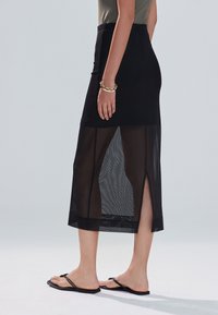 Gonna midi nera con sezione inferiore trasparente, spacchi laterali e cintura elastica. Abbinata a infradito nere e braccialetto d'oro.