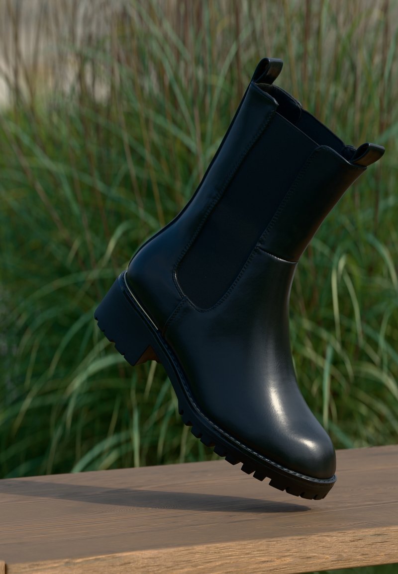 Bottine noire en cuir avec panneaux latéraux élastiques et languettes, montrée en l’air au-dessus d’une surface en bois avec un fond de feuillage vert.