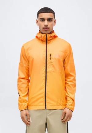 Modèle masculin de face portant une veste orange zippée avec une poche poitrine et un pantalon beige, sur un fond blanc uni.