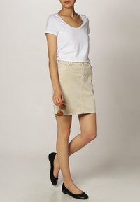 Gonna di denim beige con vestibilità dritta e tasche frontali, abbinata a un top bianco a maniche corte. Gli accessori includono occhiali da sole rotondi.