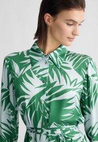 Abito camicia colletto verde e azzurro chiaro con motivo a foglie tropicali, dotato di bottoni frontali e una cintura auto-allacciata in vita.