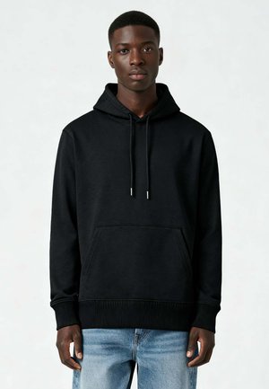 Giovane uomo con indosso una felpa nera semplice con tasca anteriore e jeans azzurri, rivolto verso l'obiettivo su uno sfondo neutro.