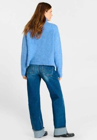 Vrouw in een lichtblauwe fuzzy trui, met brede, opgerolde blauwe jeans en zwarte schoenen, staand tegen een effen witte achtergrond.