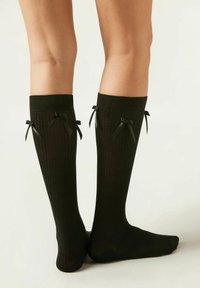 Knielange schwarze Socken mit vertikaler Rippstruktur, ausgestattet mit schwarzen Band-Schleifen an der Oberseite, aus einem weichen, strukturierten Material gefertigt.