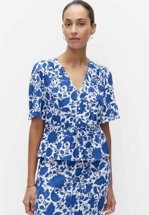 Kobieta ubrana w niebiesko-białą bluzkę w kwiaty z krótkim rękawem, zapinaną na guziki, oraz dopasowaną spódnicę, stojąca na jednolitym tle.