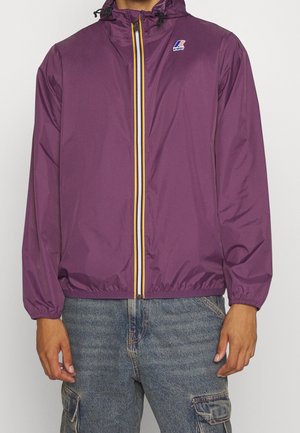 Mann trägt eine lila Kapuzenjacke mit gelben, weißen und marineblauen Reißverschlussdetails, kombiniert mit blauen Denim-Cargohosen.