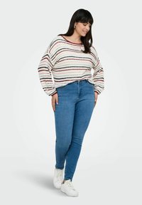 Nainen, joka pukeutunut raidalliseen neulepaitaan, sinisiin skinny-farkkuihin ja valkoisiin tennareihin, seisoo yksi jalka hieman edessä tasaisella taustalla.