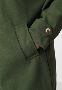 Green coat detail featuring a textured fabric, button closure, and side pocket with stitched edges for added durability.