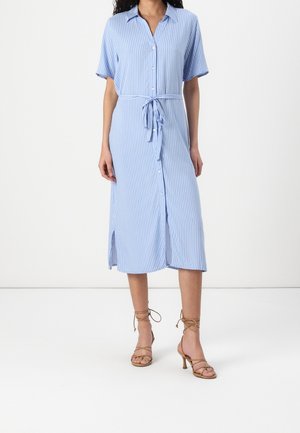 Kobieta ubrana w midi sukienkę koszulową z pionowymi, jasnoniebiesko-białymi paskami, krótkimi rękawami, guzikami z przodu, paskiem w talii oraz beżowymi sandałami na obcasie z paskami.