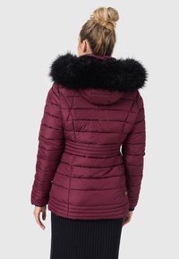 Cappotto imbottito bordeaux con cappuccio rimovibile foderato in pelliccia nera, vita sbloccata, cuciture orizzontali e un discreto logo sulla manica.