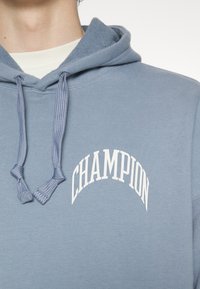 Světlemodrá mikina s kapucí s bílým logem "CHAMPION", texturovaná tkanina a nastavitelné šňůrky. Obsahuje klokánkovou kapsu a žebrované manžety.