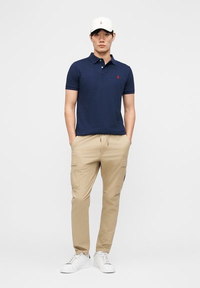 Uomo con polo blu navy, pantaloni cargo beige, scarpe da ginnastica bianche e cappellino da baseball bianco, in piedi con le mani nelle tasche.