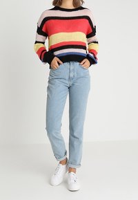 Kolorowy sweter w paski z różowymi, żółtymi i czarnymi akcentami, zestawiony z jasnoniebieskimi dżinsami i białymi sneakersami. Miękka dzianinowa tekstura.