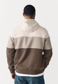 Kapuco s puloverjem v svetlo bež, temno bež in rjavi barvi z barvnim blokom. Izdelan iz mehke tkanine z narezovanimi manšetami in spodnjim robom, vsebuje kanglrovo žep.
