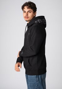 Zwarte hoodie met een trekkoord, een voorzak en bedrukte accenten op de capuchon. Draagt het met blauwe jeans.