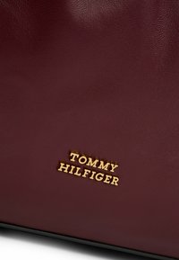 Bordeaux læderhåndtaske med et gold prægede "TOMMY HILFIGER" logo, glat tekstur og struktureret form.