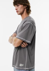 Grå oversized t-shirt med korte ærmer, synlige sømdetaljer og et logo-patch ved kanten. Stoffet ser blødt og glat ud.