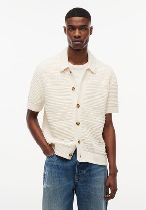 Άνδρας που φοράει ένα κρεμ κοντομάνικο πλεκτό ζακέτα με κουμπιά πάνω από ένα λευκό μπλουζάκι και μπλε τζιν, στέκεται με το ένα χέρι στην τσέπη.