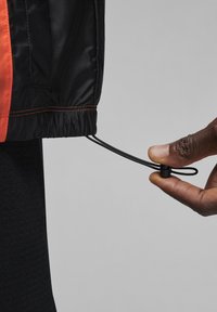 Giacca nera con accenti arancioni; presenta un cordino all'orlo, regolabile tramite un toggle. Tessuto liscio e leggero, vestibilità strutturata.