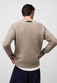 Beżowy sweter wykonany na drutach, o miękkiej teksturze, z prążkowanymi mankietami i brzegiem oraz prostokątną czarną skórzaną metką z tyłu. Zaprojektowany w luźnym kroju.