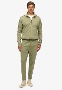 Olijfgroene trainingsoutfit van zachte stof; jasje met ritssluiting en zwarte strepen, bijpassende broek met een trekkoord in de taille, gestyled met beige sneakers.