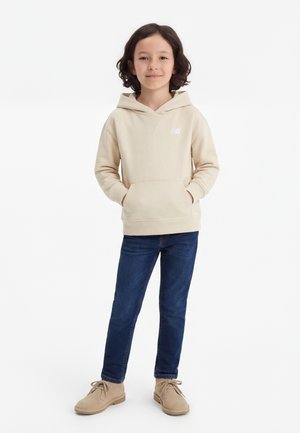 Joven de pie con las manos en los bolsillos, vistiendo una sudadera con capucha beige, jeans azules y zapatos marrón claro con cordones, sobre un fondo blanco liso.