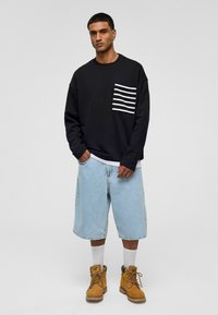 Zwarte sweatshirt met witte horizontale strepen op de borstzak, gecombineerd met lichtblauwe denim shorts en bruine werklaarzen.