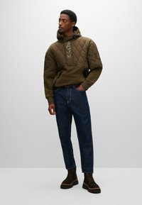 Quiltet olivengrønn hettegenser med frontglidelåser, kombinert med mørkeblå denimjeans og brune snøresko. Teksturert materiale og design detaljer.
