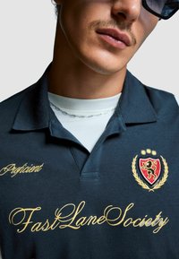 Marineblå pikétröja i bomull med guld broderad text "Proficient" och "FastLaneSociety", samt en grevskapssymbol och lagerkransdesign.