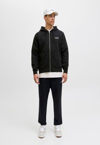 Schwarze zip-up Kapuzenjacke mit weißem Logo auf der Brust, kombiniert mit schwarzen Sweatpants und weißen Turnschuhen. Das Modell trägt außerdem eine weiße Mütze. Leichtes Material.