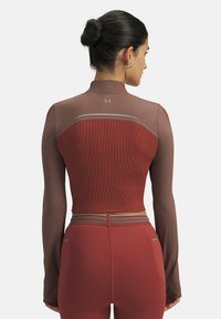 Kvinde med hår i en knold iført tætsiddende langærmet crop-top og matchende højtaljede leggings i dæmpede røde nuancer, vendt væk.