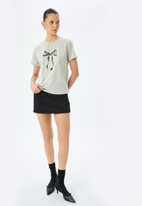 Grå t-shirt med en sort sløjfe grafik, parret med en sort miniskørt. Modellen bærer sorte sokker og spidse patenterede sko.