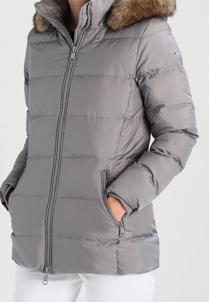 Veste d'hiver matelassée grise avec capuche bordée de fourrure, fermeture éclair frontale, poches latérales zippées, portée par une personne avec les mains dans les poches et un pantalon blanc.