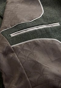 Chaqueta de tela gris con un detalle texturizado y estampado, con secciones verdes en contraste y ribetes beige a lo largo de los bolsillos.