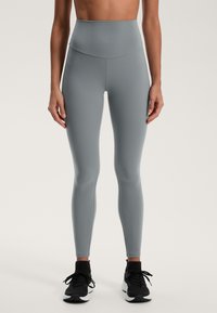 Svetlošedé legíny s vysokým pásom vyrobené z elastického materiálu, s hladkou textúrou a bezšvovým dizajnom, v kombinácii s čiernymi športovými topánkami.