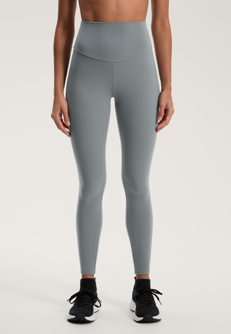 Svetlošedé legíny s vysokým pásom vyrobené z elastického materiálu, s hladkou textúrou a bezšvovým dizajnom, v kombinácii s čiernymi športovými topánkami.