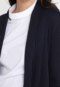 Žena v bílé halence s vysokým límcem pod tmavě modrým žebrovaným kardiganem, s viditelnými rameny a horní částí trupu.