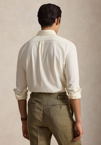 Mees, kes kannab kreemjat pikkade varrukatega särki, varrukad üles keritud, ja pruunid kohandatud püksid, seisab neutraalse tausta ees seljaga vaataja poole.