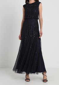 Vestido de noche sin mangas en azul marino, con un cuerpo fruncido, adornado con lentejuelas y una falda de tul fluida. Sandalias negras con tiras.