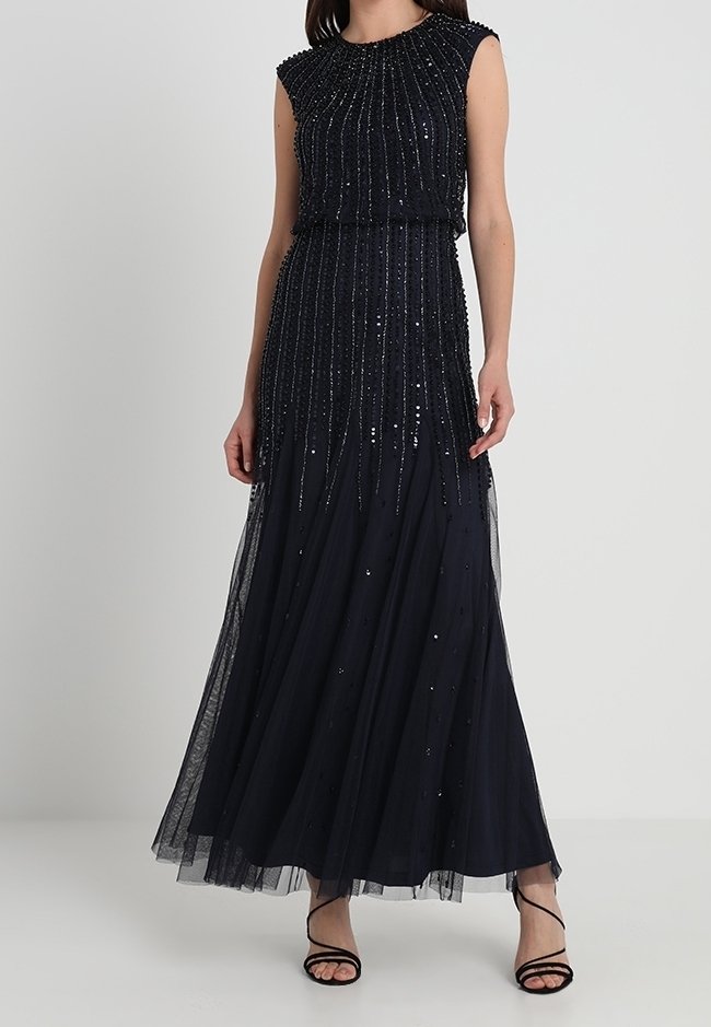 Vestido de noche sin mangas en azul marino, con un cuerpo fruncido, adornado con lentejuelas y una falda de tul fluida. Sandalias negras con tiras.