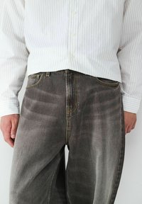 Weißes Hemd mit Knöpfen und vertikalen Streifen, kombiniert mit grauen, ausgewaschenen Jeans mit gelber Naht und weitem Schnitt.