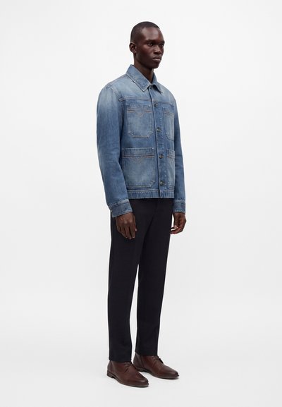 Homme portant une veste en jean bleue, un pantalon noir et des chaussures habillées marron, debout devant un fond blanc uni.