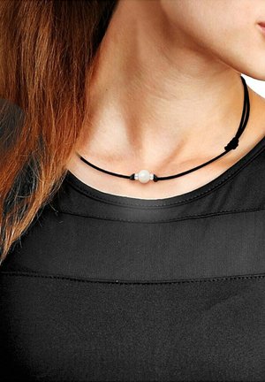 Schwarze Kordel-Halskette mit einer zentralen weißen Perle, flankiert von kleinen metallischen Akzenten, getragen über einem schwarzen Oberteil mit einem strukturierten Design.